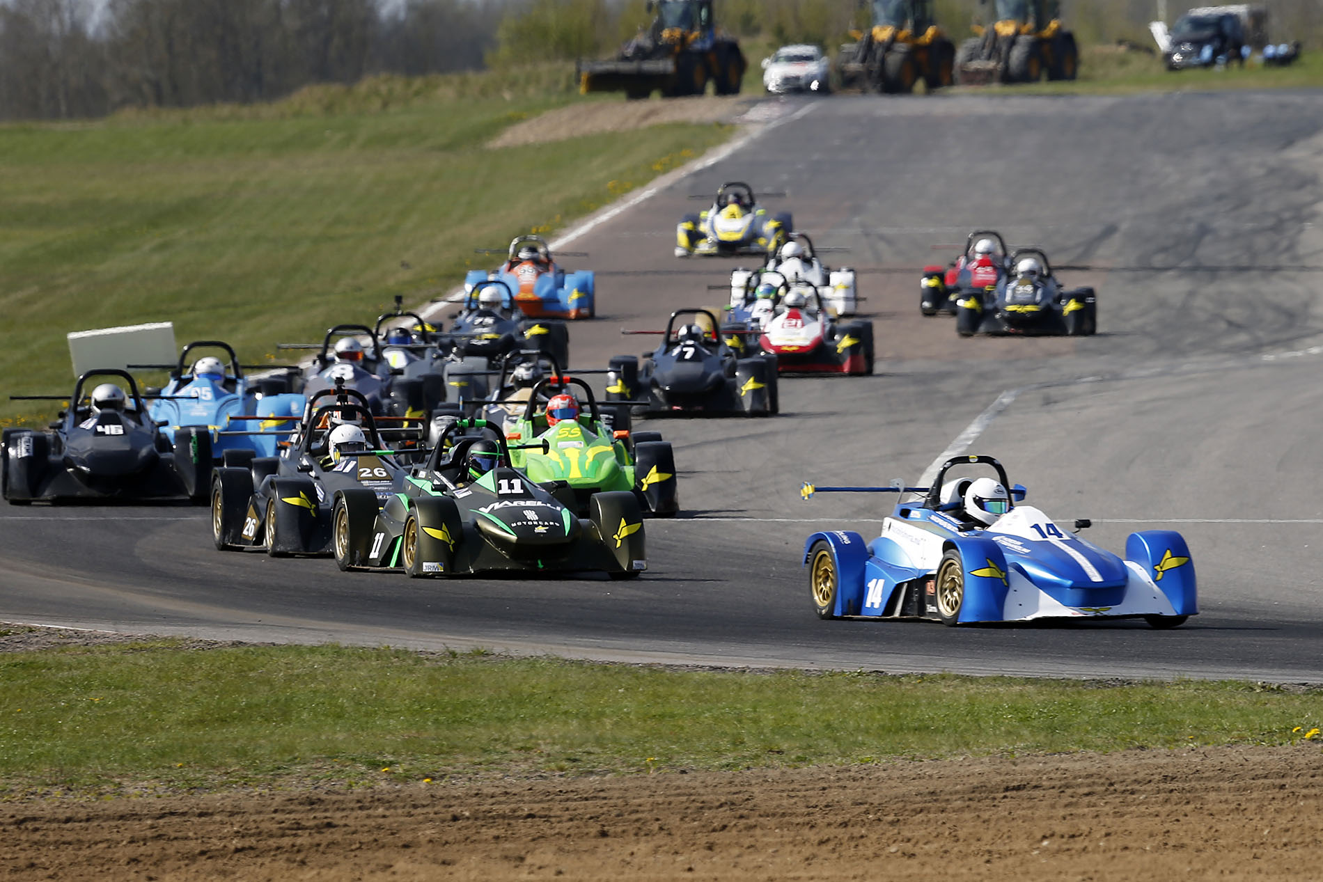 A group of race cars fills the frame, with drivers wearing white helmets. The race begins on the straightaway, followed by...