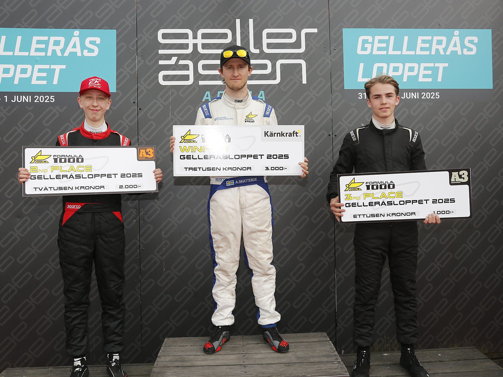 The image shows three men in racing suits holding signs at a podium. * The two on the outside are in black, and the one in...