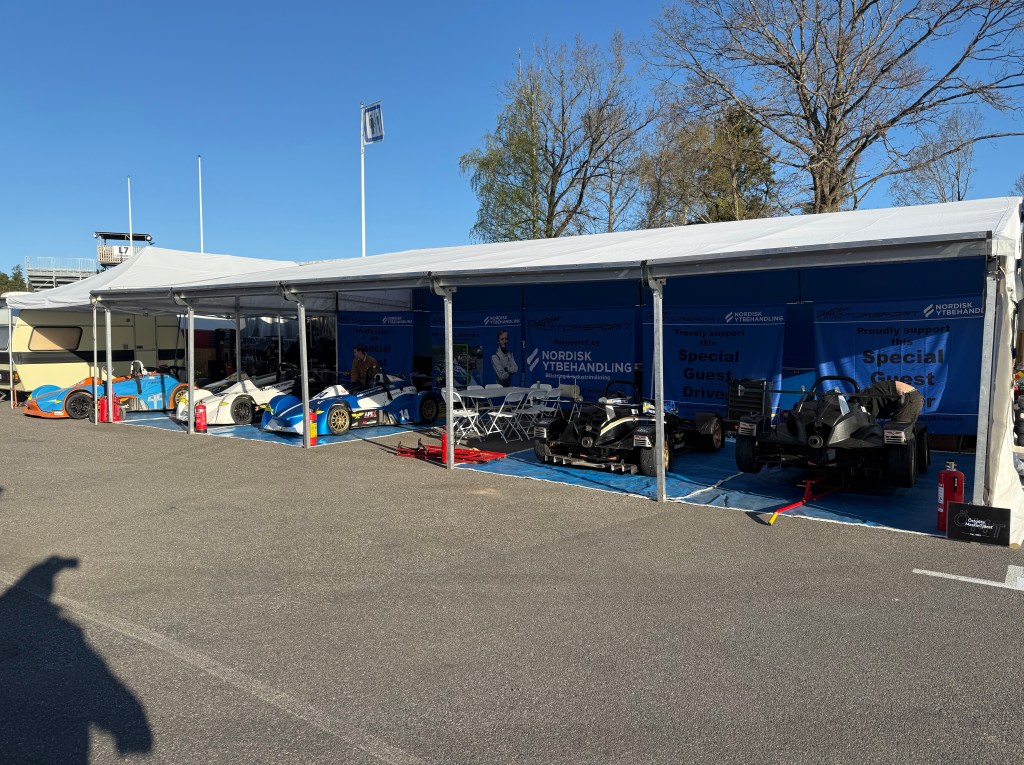 **Summary:** A photograph of a row of race cars and support vehicles under a white canopy. **Foreground:** In the left hal...